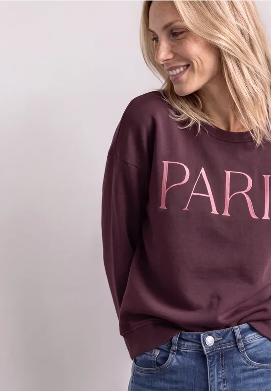 Glunderende vrouw in jeans en bordeauxroze trui met 'PARIS' op een eenvoudige achtergrond.