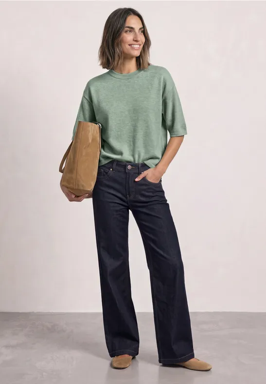 Frau trägt grünes T-Shirt und weite dunkle Jeans, hält eine braune Tasche und lächelt.
