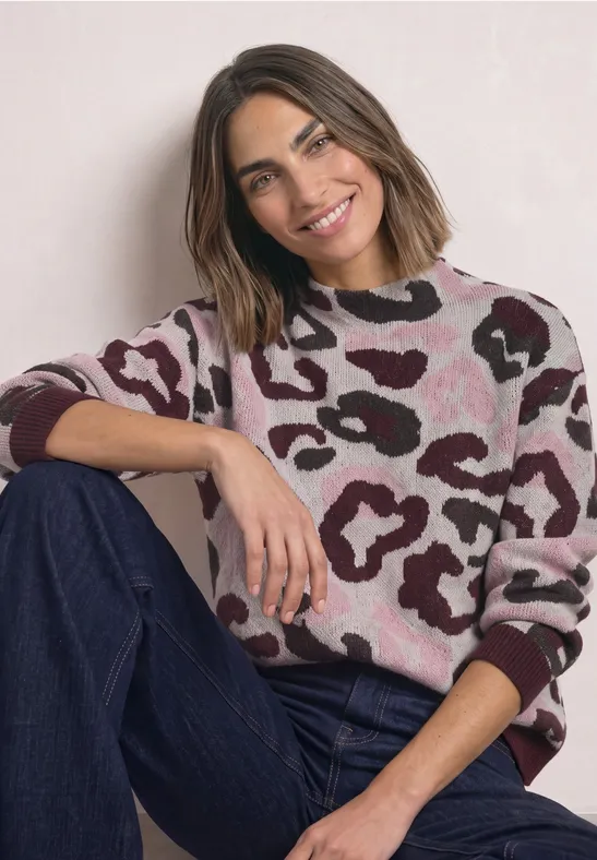 Eine lächelnde Frau sitzt entspannt, trägt einen rosa-braunen Pullover mit Muster und eine blaue Jeans.