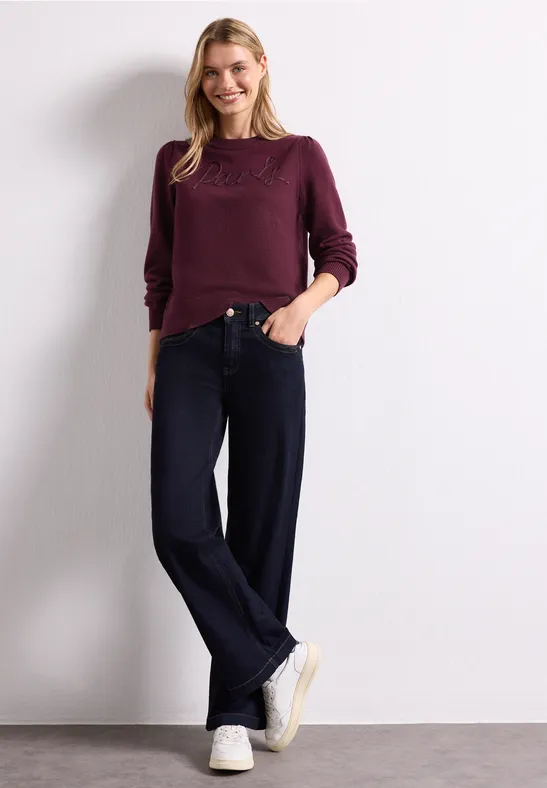 Une femme en pull bordeaux, jean large et baskets blanches sur fond blanc