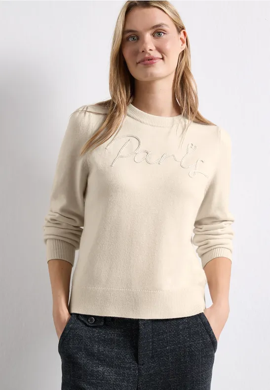 Femme portant un pull crème 'Paris' et un pantalon à carreaux gris, mains dans les poches.