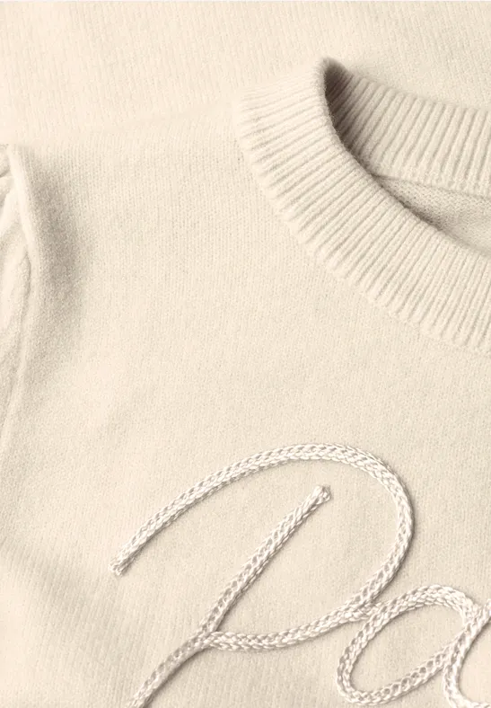 Gros plan sur un pull crème à col côtelé avec broderie cursive argentée