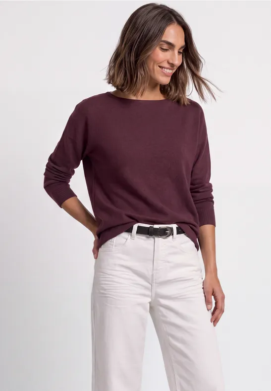 Vrouw lacht in een bordeauxrode trui en lichte jeans, ontspannen houding.