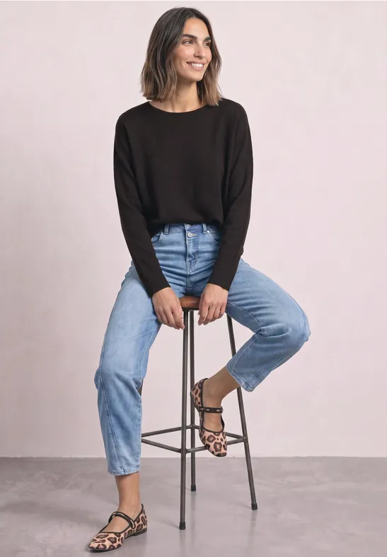Vrouw lacht, zit op kruk, draagt zwarte trui, blauwe jeans en luipaardprint sandalen.