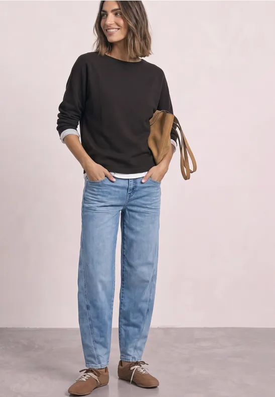 Vrouw in zwarte trui, wit overhemd, lichtblauwe jeans en bruine loafers voor een lichte achtergrond.