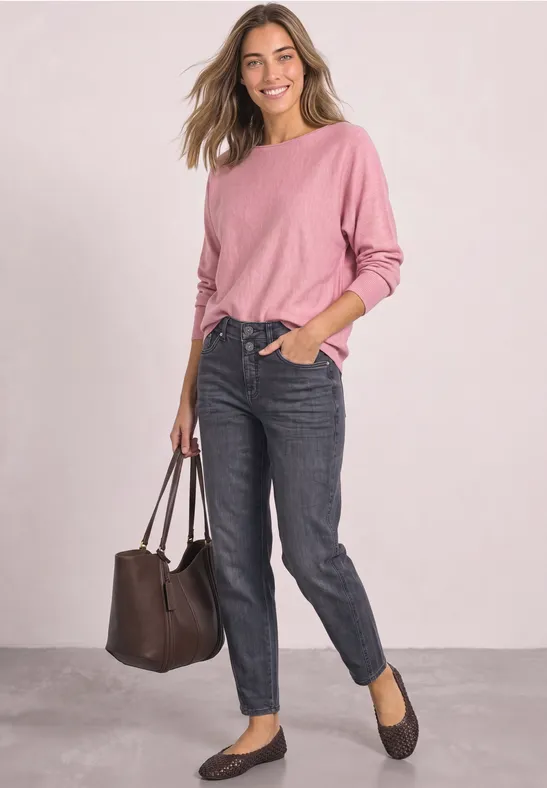 Frau in rosa Pullover und dunklen Jeans mit brauner Tasche und schwarzen Ballerinas vor hellem Hintergrund.