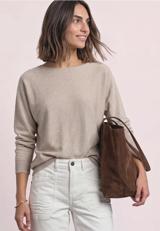 Vrouw met halflang haar in beige trui en witte jeans, met een bruine suède tas over de schouder.