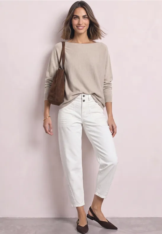 Een vrouw draagt een beige trui, een witte broek, een bruine schoudertas en donkere ballerina’s voor een lichte muur.