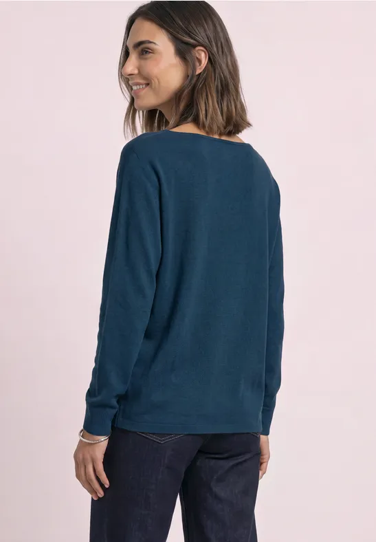 Frau im dunkelblauen Pullover und Jeans, seitlich stehend, Blick über die Schulter vor rosa Hintergrund.