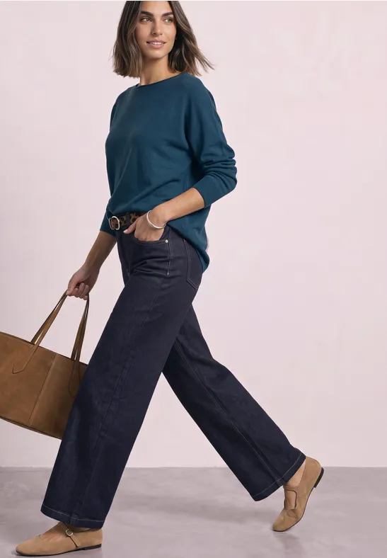Frau in weitem Jeans, blaugrünem Pullover und brauner Tasche, vor rosa Hintergrund, lässig gehend.