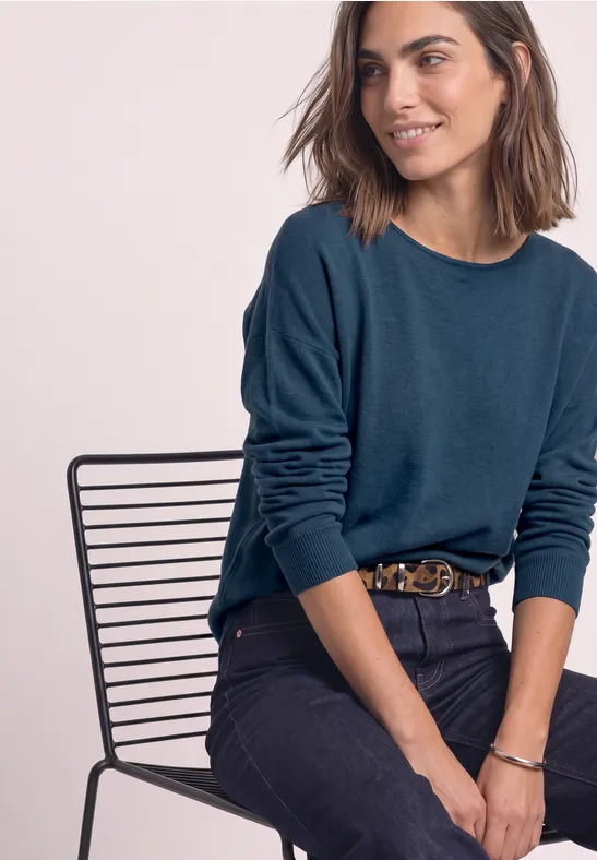 Frau sitzt lächelnd auf einem Drahtstuhl, trägt blauen Pullover und dunkle Jeans.