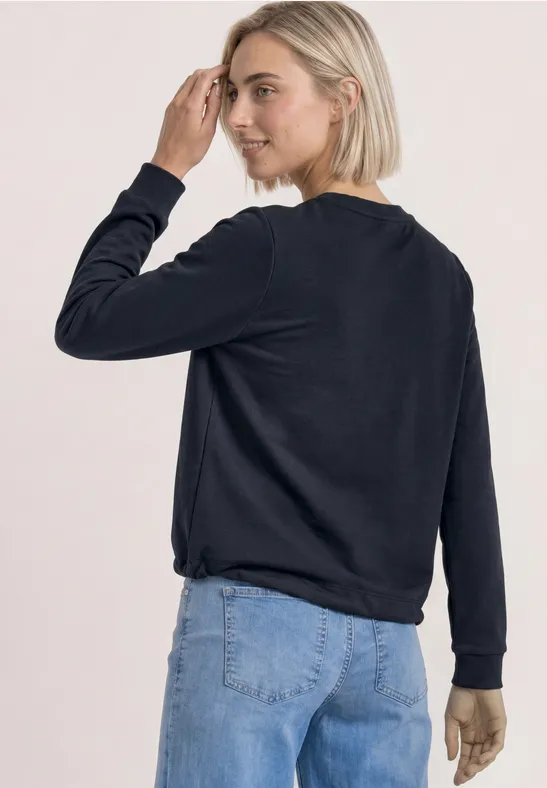 Blonde Frau in schwarzem Pullover und Jeans, seitliche Rückenansicht.
