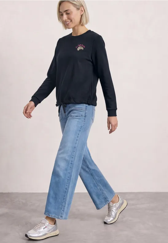 Frau in schwarzem Langarmshirt, weitem Jeanshose und silbernen Sneakers vor hellem Hintergrund.