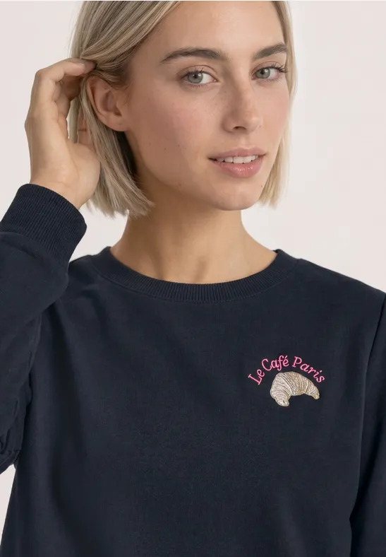 Frau mit kurzem blondem Haar in dunkelblauem Pullover, bestickt mit Croissant und Schriftzug „Le Café Paris“.