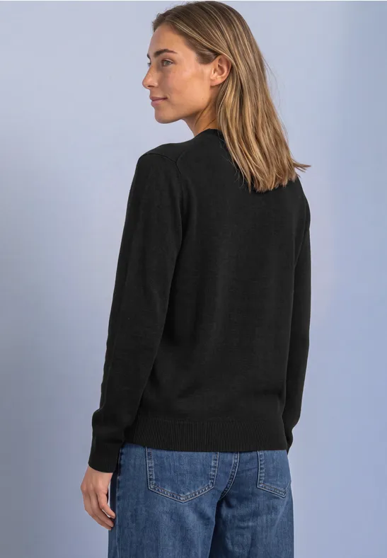 Frau steht seitlich vor hellem Hintergrund, trägt schwarzen Pullover und blaue Jeans.