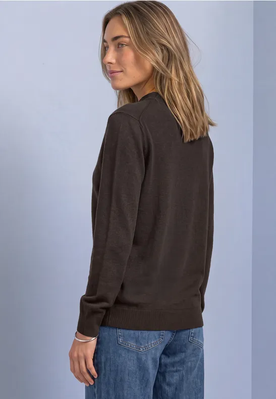 Frau in dunklem Pullover und blauen Jeans steht seitlich vor hellem Hintergrund.