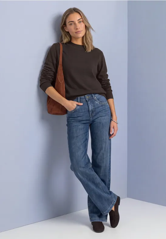 Eine Frau lehnt entspannt an lilafarbener Wand, trägt schwarzen Pullover, blaue Jeans und braune Schultertasche.