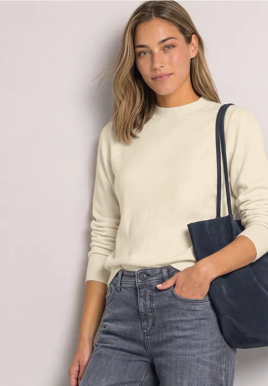 Frau in Jeans und beigem Pullover mit schwarzer Schultertasche und einer Hand in der Hosentasche.
