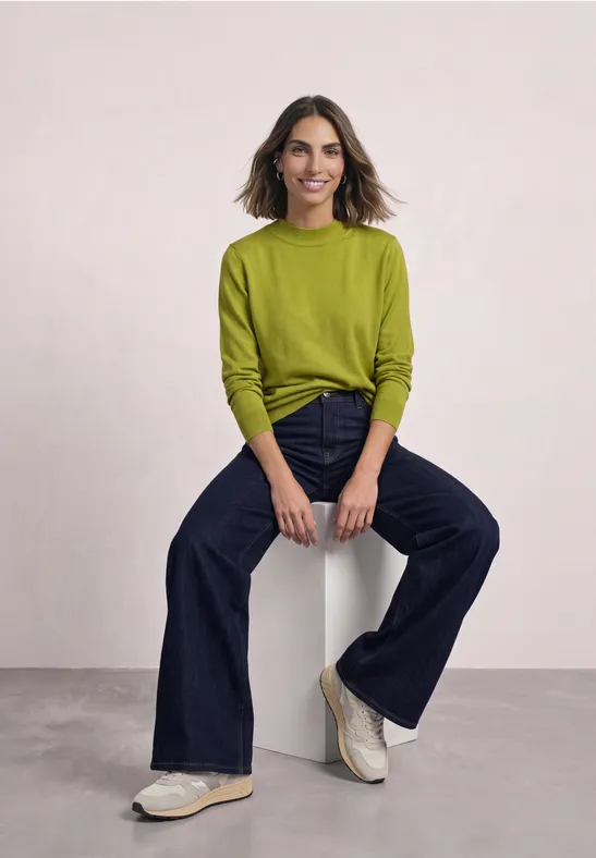 Eine lächelnde Frau sitzt auf einem Hocker, trägt ein grünes Oberteil, dunkle Jeans und weiße Sneaker.