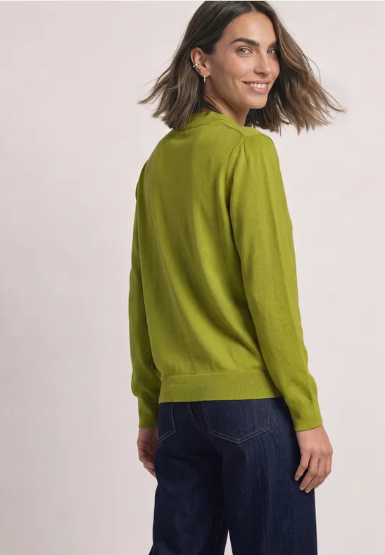 Eine Frau in grüner Strickjacke und Jeans schaut lächelnd über die Schulter.