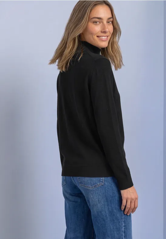 Junge Frau mit schwarzem Rollkragenpullover und blauen Jeans, schaut lächelnd über die Schulter vor hellem Hintergrund.
