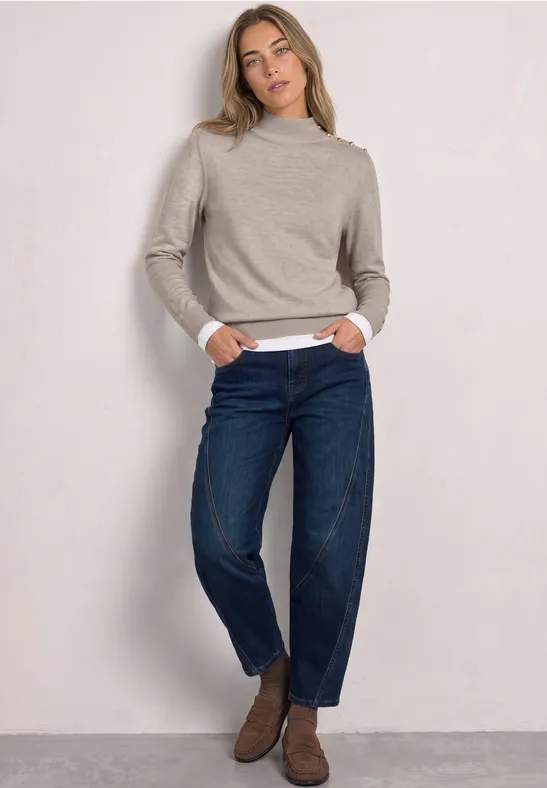 Frau in beige Rollkragenpullover, weißem Shirt und dunkelblauen Jeans steht lässig vor hellem Hintergrund.
