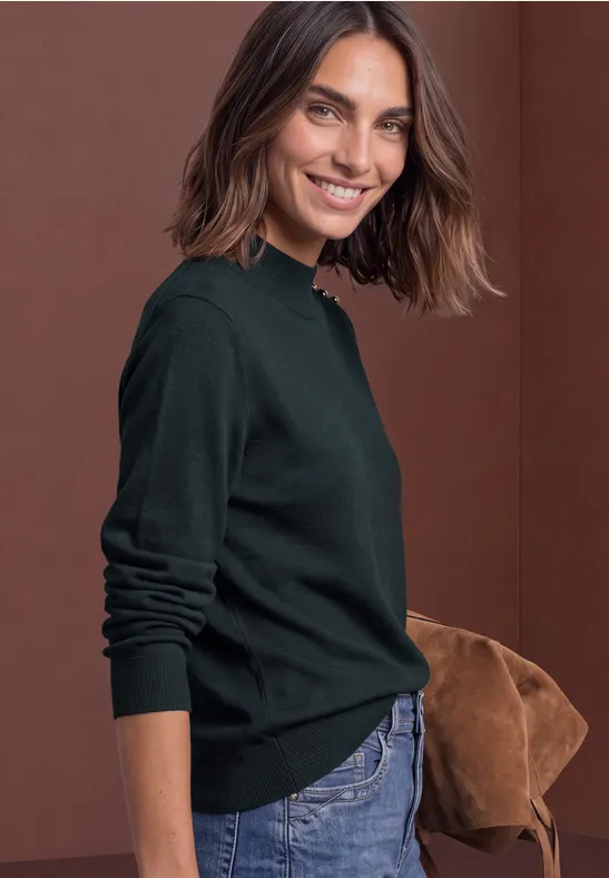 Lächelnde Frau in grünem Pullover und Jeans mit brauner Tasche vor rotem Hintergrund.
