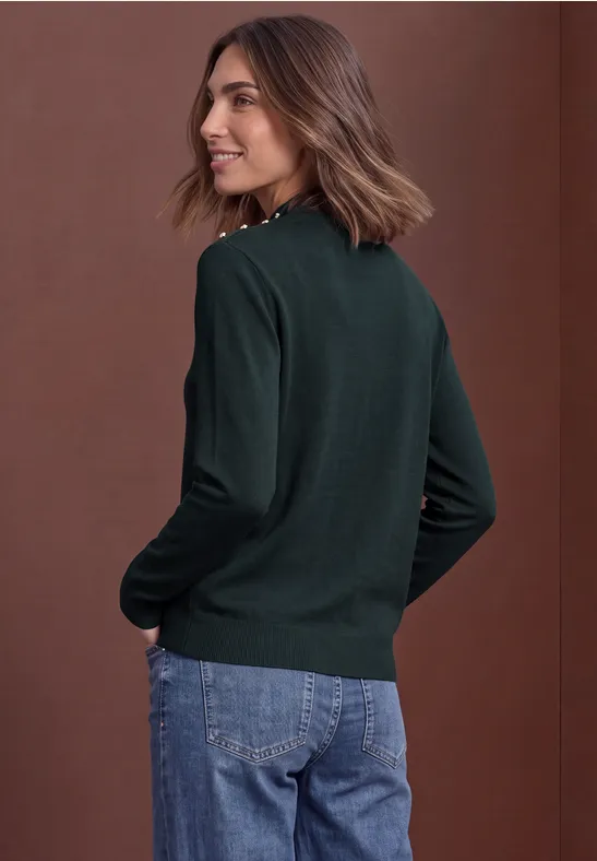 Frau im dunkelgrünen Pullover und Jeans, im Profil stehend, vor braunem Hintergrund.