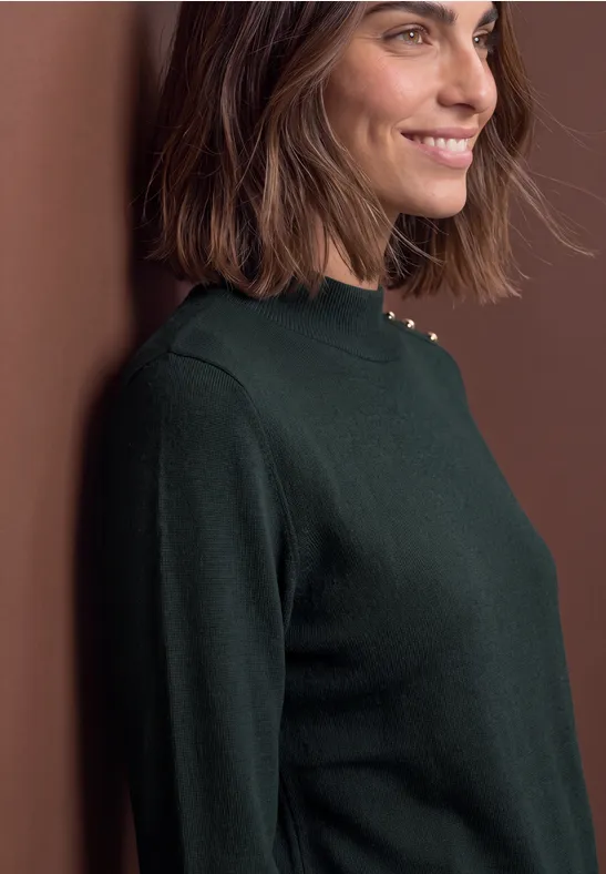 Eine lächelnde Frau lehnt seitlich an einer Wand und trägt einen dunkelgrünen Pullover.