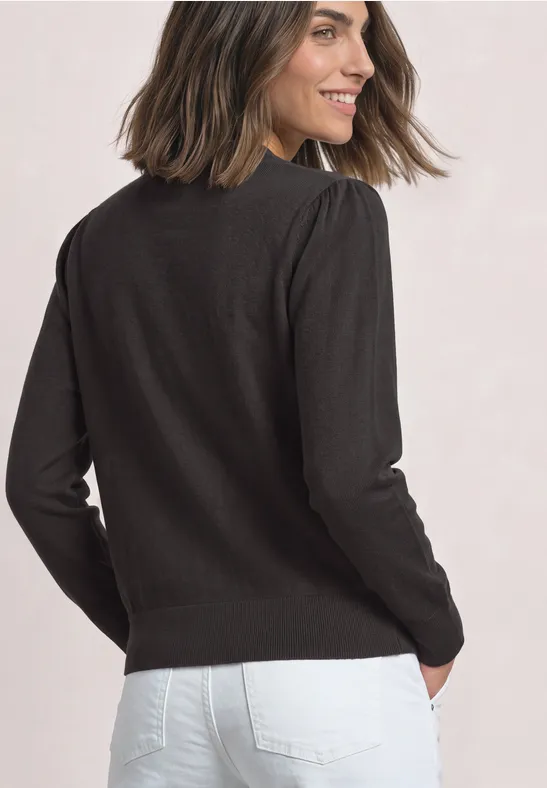Frau in schwarzem Pullover und weißer Jeans, von hinten mit Blick über die Schulter, lächelnd.