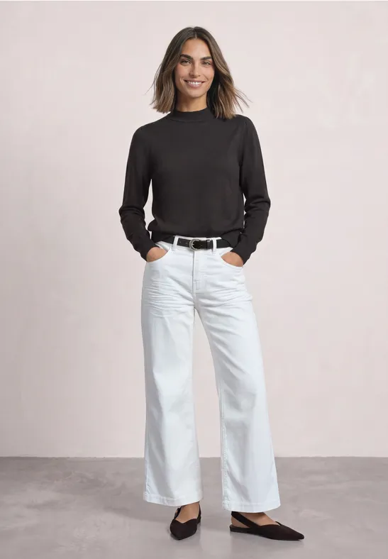 Frau in weitem weißen Jeans, schwarzem Oberteil, Gürtel und Ballerinas, lächelnd vor neutralem Hintergrund.