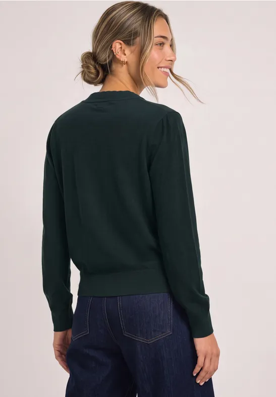 Eine Frau trägt einen dunkelgrünen Pullover und Jeans, schaut lächelnd über die Schulter.