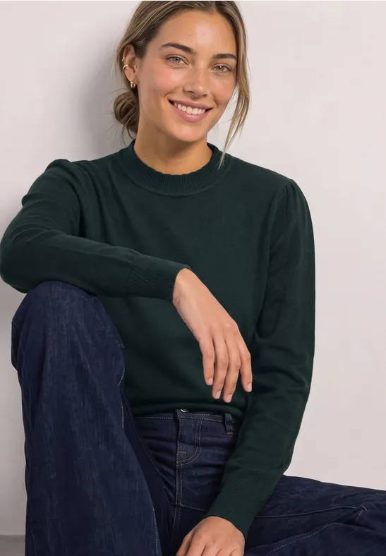 Frau sitzt lächelnd, trägt dunkelgrünen Pullover und blaue Jeans vor hellem Hintergrund.