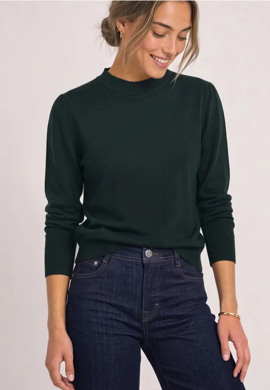Frau im dunkelgrünen Pullover und Jeans lächelt und schaut nach unten.
