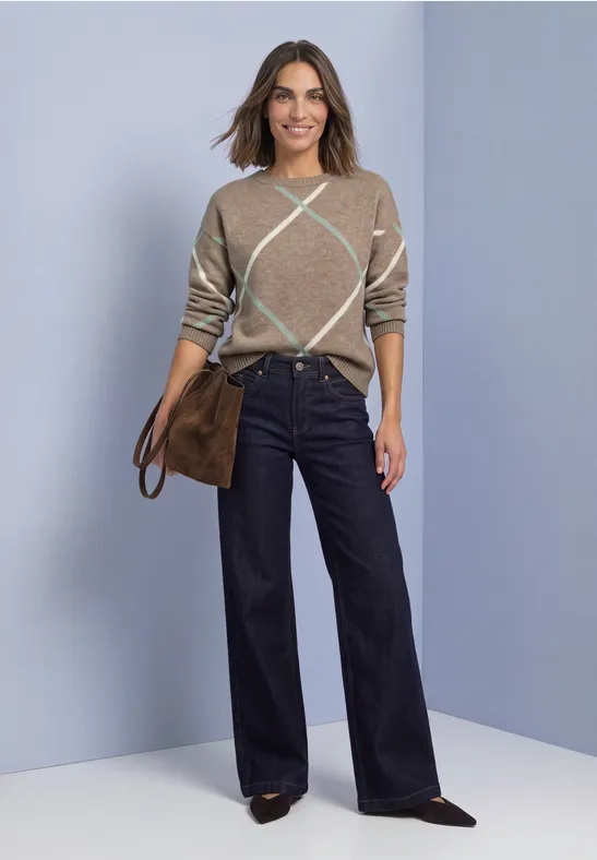 Frau in beige-kariertem Pullover, weiten dunkelblauen Hosen und brauner Tasche vor hellem Hintergrund.