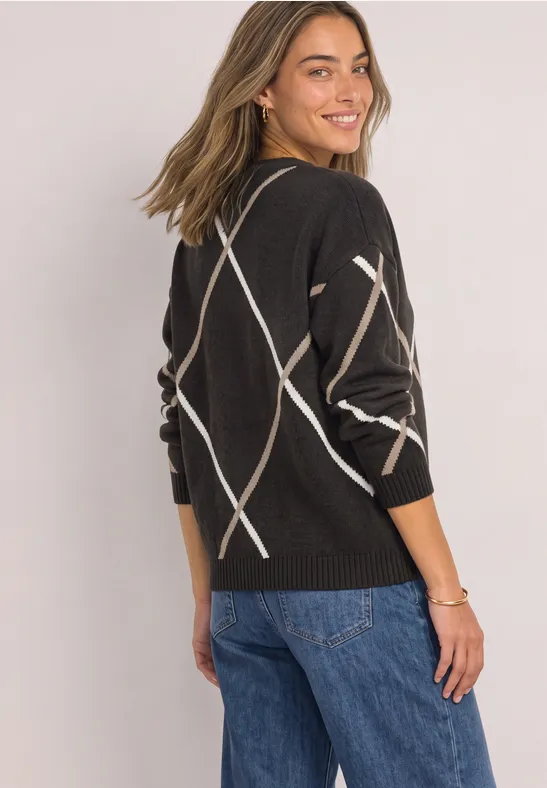 Eine Frau in schwarzem Pullover mit beigen Rautenmustern und Jeans lächelt über die Schulter.