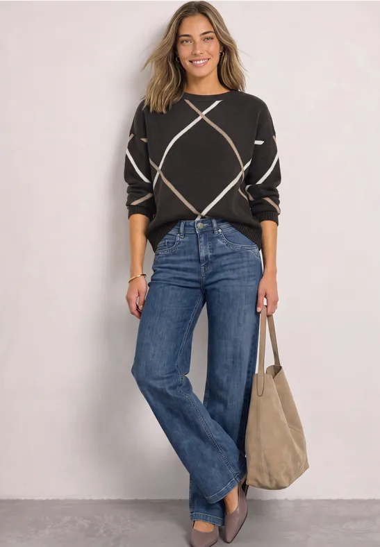 Frau in schwarzem Pullover mit Rautenmuster, blauen Jeans und beiger Tasche, lächelnd vor heller Wand.