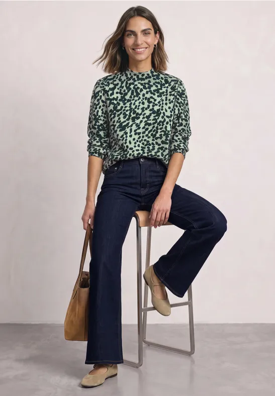 Vrouw in groen dessinblouse en donker spijkerbroek zit ontspannen op een hoge kruk, glimlachend.