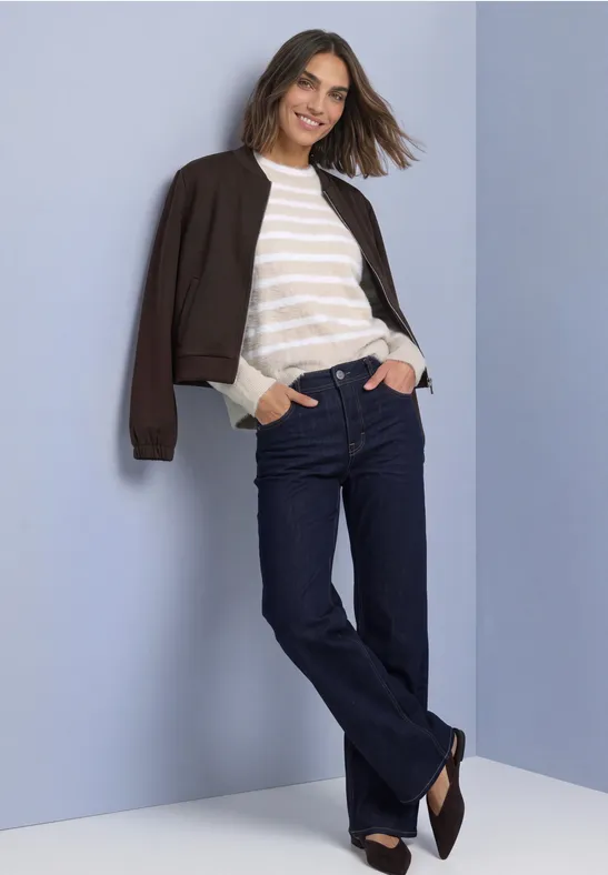 Eine lächelnde Frau lehnt entspannt an einer Wand, trägt Jeans, gestreiftes Shirt und dunkle Jacke.