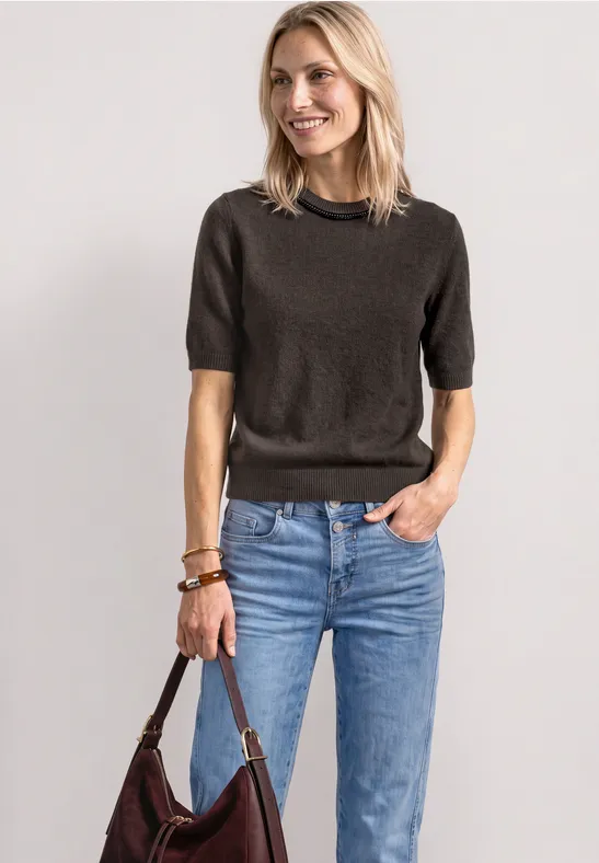 Lächelnde Frau in dunklem Kurzarmpullover, blauen Jeans und brauner Ledertasche.