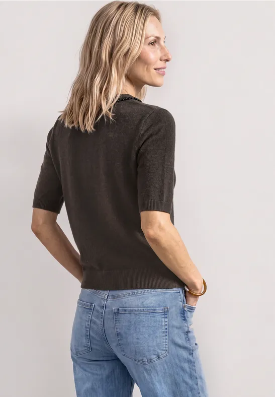 Eine Frau in schlichter, dunkelbrauner Strickbluse mit halblangen Ärmeln und blauen Jeans vor hellem Hintergrund.
