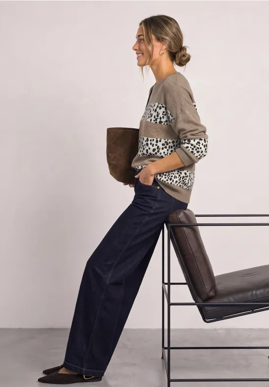 Femme en gilet à motif et jean large appuyée sur une chaise, tenant une veste.