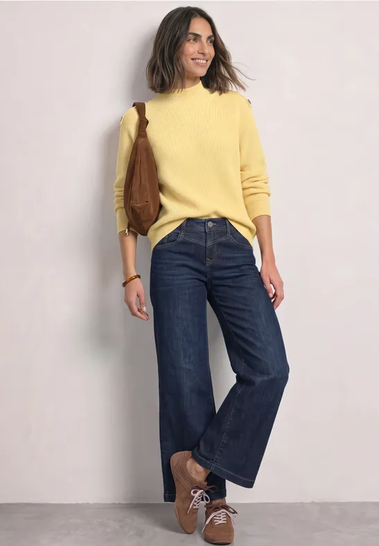 Frau lehnt an Wand, trägt gelben Pullover, weite Jeans, braune Schuhe und eine braune Tasche.