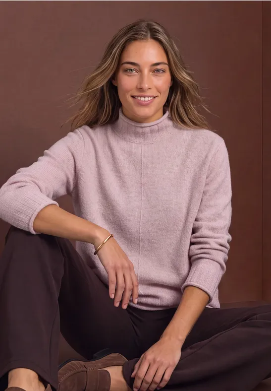 Eine lächelnde Frau sitzt entspannt auf dem Boden, trägt einen rosa Pullover und braune Hosen vor neutralem Hintergrund.