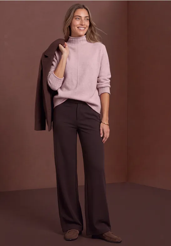 Stilvolle Frau in rosa Pullover und weiten braunen Hosen trägt Blazer lässig über der Schulter.