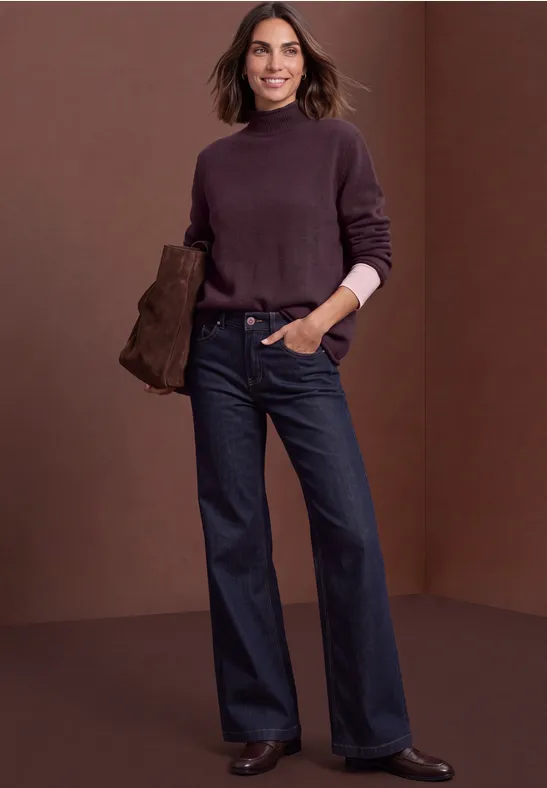 Frau in dunkelviolettem Rollkragenpullover und weiten Jeans hält eine Clutch vor braunem Hintergrund.