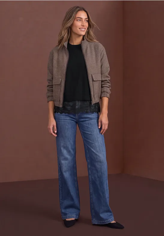 Frau in brauner Jacke, schwarzem Shirt und weiten Jeans vor braunem Hintergrund.