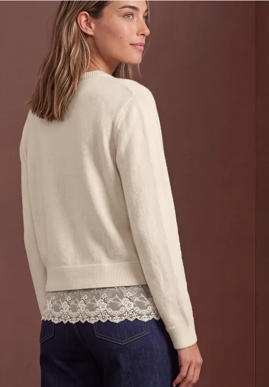 Frau trägt beigefarbene Strickjacke mit Spitzen-Top und Jeans, Blick über die Schulter vor braunem Hintergrund.