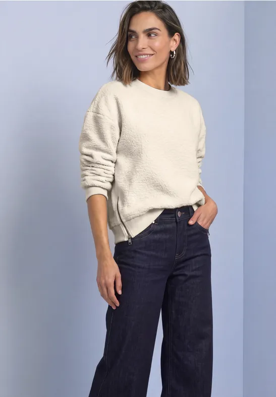 Eine Frau trägt einen hellen Pullover und dunkle Jeans vor neutralem Hintergrund.