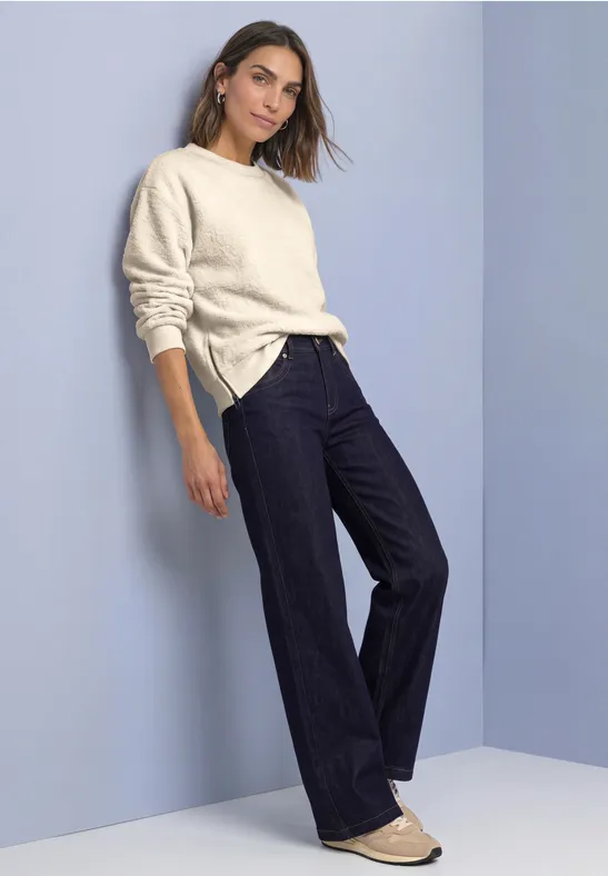 Frau lehnt an Wand, trägt cremefarbenen Pullover, dunkle Schlagjeans und helle Ballerinas vor schlichtem Hintergrund.